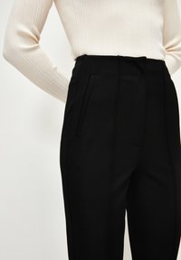 Black tailored trousers with a smooth texture, featuring side pockets and a high waist design. Worn with a light beige ribbed sweater.