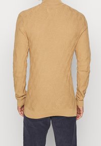 Strickpullover in hellem Beige mit strukturiertem Rautenmuster, ausgestattet mit langen Ärmeln und hohem Kragen, kombiniert mit dunkler Hose.