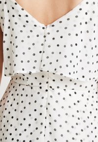 Blusa blanca con lunares negros, diseño superpuesto, escote redondeado y cierre de botón en la parte posterior; la tela tiene una textura suave y ligera.