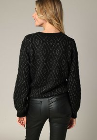 Femme de dos portant un pull noir à motifs et un pantalon en cuir noir sur fond uni.