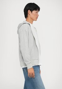 Sudadera gris con cremallera y capucha ajustable, puños de canalé y bolsillos frontales, llevada sobre una camiseta blanca, combinada con jeans de mezclilla azules.