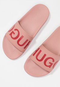 Růžové gumové pantofle s texturovanou stélkou a širokými pásky s výrazným červeným nápisem "HUGO". Bez podpatku, otevřená špička.