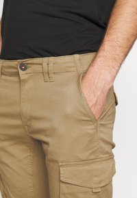 Béžové cargo kalhoty s bočními kapsami, kontrastním šitím a knoflíkovým zapínáním v pase. Tkanina má hladký povrch.