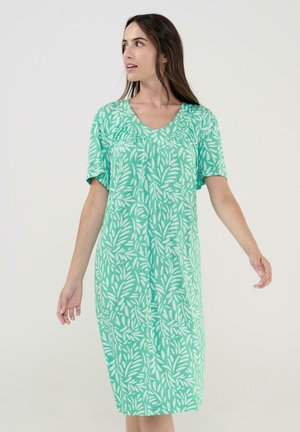 Mujer con vestido hasta la rodilla color verde azulado con patrón de hojas blancas, mangas cortas y escote en V, de pie con los brazos ligeramente extendidos, mirando hacia la izquierda.