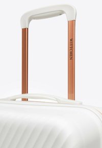Maleta blanca con textura acanalada, que presenta un asa extensible de color cobre y un asa blanca en la parte superior. El nombre de la marca "WITTCHEN" es visible.