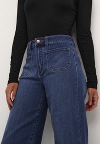 Vaqueros azules de mezclilla de talle alto con pernera recta, cinco bolsillos y un botón de cobre. La tela muestra una textura suave con un diseño clásico.