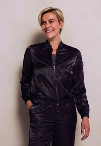 Schwarze Satin-Bomberjacke mit Reißverschluss, zwei Vordertaschen, gerippten Bündchen und passenden Hosen mit einer dezenten gemusterten Textur.