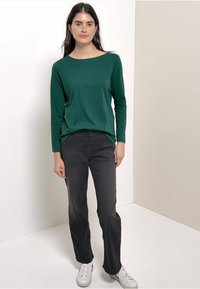 Green long-sleeve top with a wide neckline, paired with dark gray straight-leg pants and white sneakers, set against a neutral background.