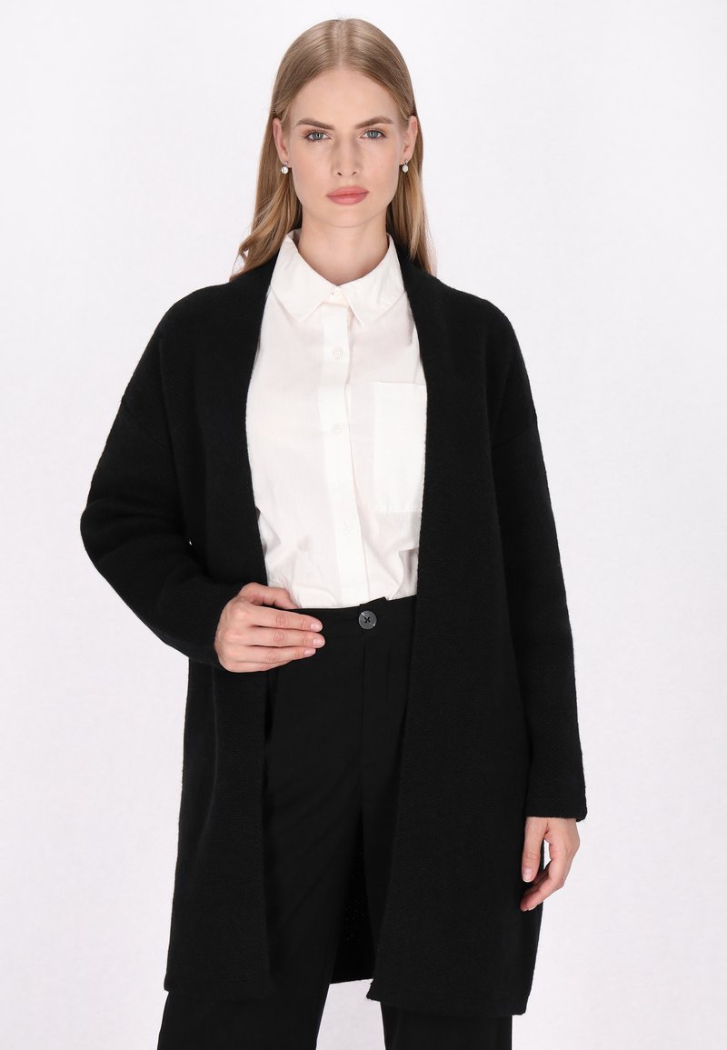 Langer schwarzer Cardigan aus weichem Stoff, mit offenem Vorderdesign, langen Ärmeln und lockerer Passform, kombiniert mit einer weißen Knopfleiste und schwarzen Hosen.