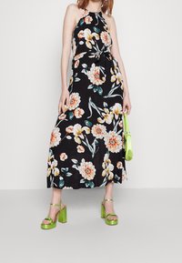 Robe à col licou à motif floral sur fond noir, ornée de fleurs orange et jaunes. Assortie à des sandales à talons carrés vert vif.