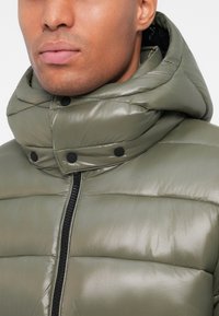 Olivenfarvet polstret jakke med høj krave, sorte trykknapper og en frontlynlås. Stoffet er skinnende og quiltet med vandrette linjer.
