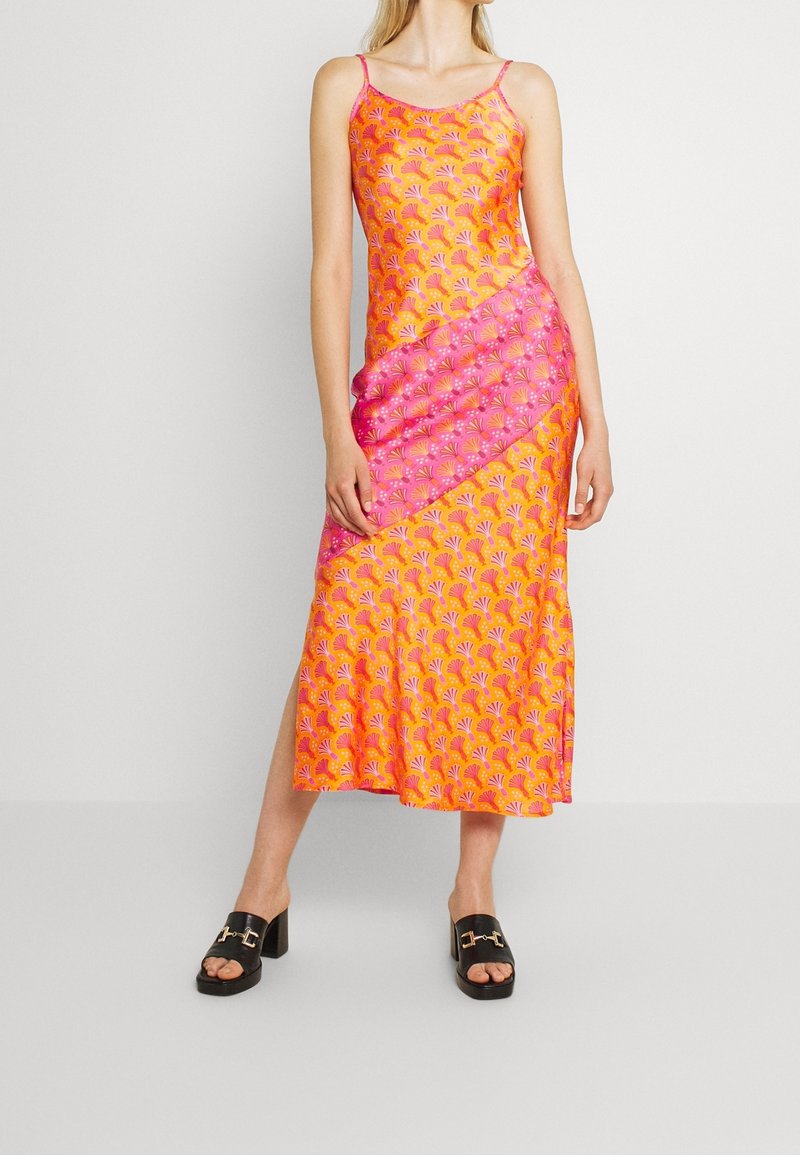Femme portant une robe midi sans manches orange et rose à motifs avec des fines bretelles et des sandales à talons noires, sur un fond uni.