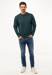 Tmavě zelená mikina s dlouhými rukávy a kulatým výstřihem, kombinovaná s modrými džíny a bílými teniskami. Hladká textura a ležérní design.