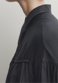 Zwarte shirt met een kraag en gathered textuur, met een ontspannen pasvorm. De stof lijkt glad met subtiele geweven details.
