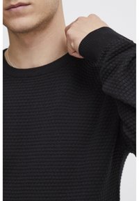 Pulover negru din tricot texturat, cu guler rotund și manșete ribbate, parțial vizibil pe o persoană. Prim-planul evidențiază modelul materialului.
