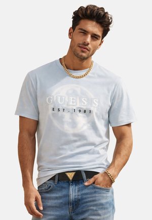 Hombre con camiseta azul claro "GUESS EST. 1981", jeans azules con cinturón negro, cadena de oro y pulsera de oro, posando con una mano en el bolsillo.