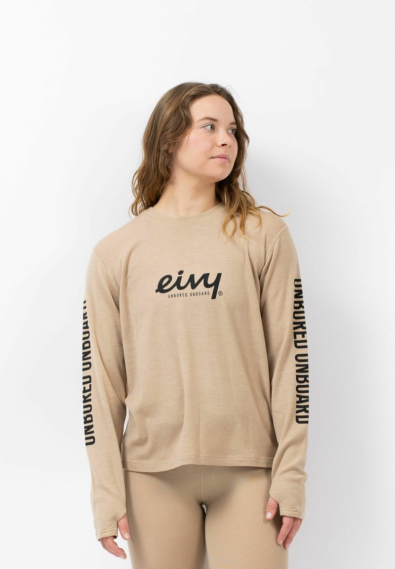 Beige langarmpolit met ribbeltextuur; beschikt over een zwart logo op de borst en zwarte tekst die verticaal op beide mouwen is gedrukt.