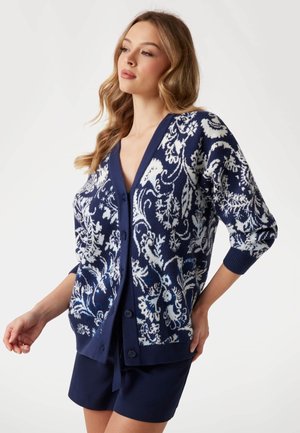 Navy cardigan with white floral pattern, v-neckline, and three-quarter sleeves. Features buttons down the front and ribbed cuffs.