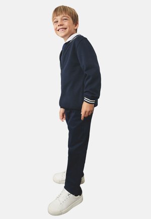 Giacca a zip marina con accenti a strisce bianche, abbinata a pantaloni coordinati e sneakers bianche, progettata per bambini. Tessuto morbido.