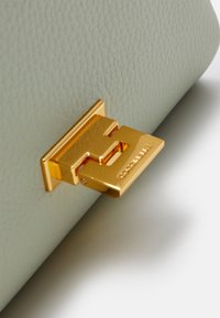 Vihreä nahkainen käsilaukku, jossa on tekstuuripinta, kullanvärinen metallilukko kohokuvioidulla brändin nimellä ja logolla.