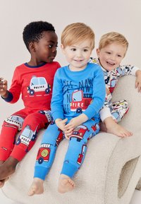 Trois enfants portent des pyjamas colorés ornés de graphiques de véhicules : des voitures de police rouges, des camions de pompiers bleus et des motifs blancs sur une surface douce et texturée.