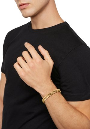 Goldfarbener Kettenarmreif mit ineinandergreifenden Gliedern, getragen am Handgelenk, vor einem schwarzen T-Shirt-Hintergrund. Glatte Textur, glänzende Oberfläche.