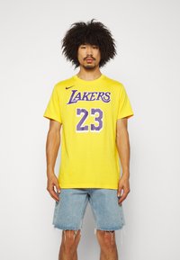 Gul kortermet T-skjorte med "LAKERS" i lilla, nummer "23" i hvitt, og Nike-logo. Brukt sammen med lyse denimshorts.