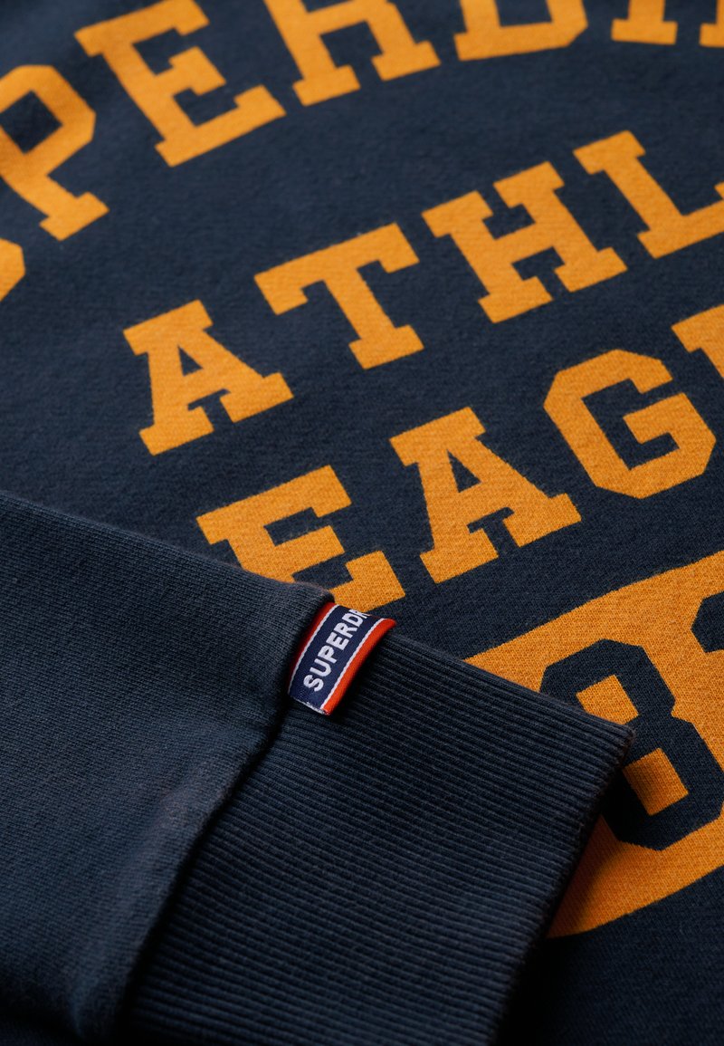 Marineblaue Sweatshirt mit auffälligem orangem Text, gerippten Bündchen und einem Markenetikett in rot und blau am Ärmel.