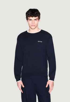 Giovane uomo con capelli ricci che indossa un maglione nero Armani Exchange e pantaloni neri, in piedi davanti a uno sfondo chiaro e uniforme.