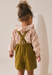 Rosa Bluse mit floralem Muster und Rüschenschild, kombiniert mit olivgrünen Cordoverall, der über kreuzliegende Träger und einen gerafften Bund verfügt.