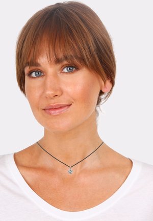 Silver pendant necklace on a thin black chain, featuring a circular charm with intricate detailing, worn with a white t-shirt.