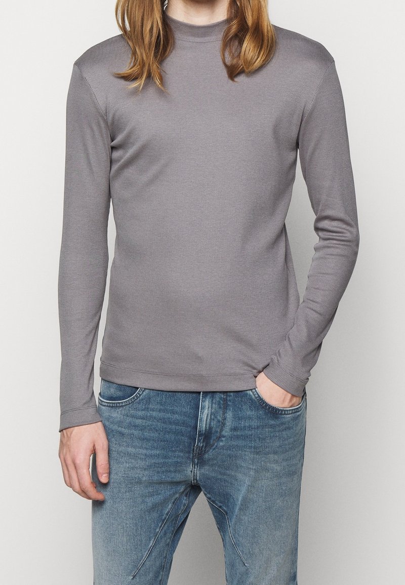 Jeune personne portant un col roulé gris à manches longues et un jean bleu, une main dans la poche, devant un fond uni.