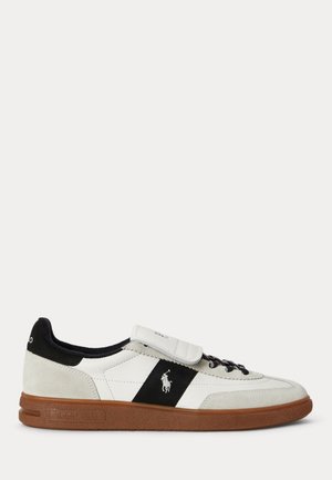 Sneaker blanc basse avec bande noire, semelle marron et logo brodé sur le côté, avec talon bordé de noir et bride velcro sur les lacets.