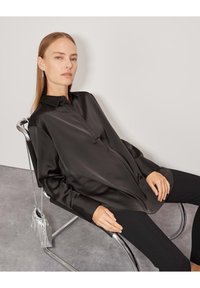 Blusa di raso nera con colletto e pattina frontale, dotata di maniche lunghe e vestibilità rilassata. La modella indossa pantaloni neri e si siede su una sedia in cromo.