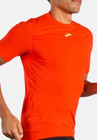 Sportovní tričko s krátkým rukávem v jasně oranžové barvě, vyrobené z lehkého, prodyšného materiálu. Obsahuje decentní logo na hrudi a přiléhavý střih.