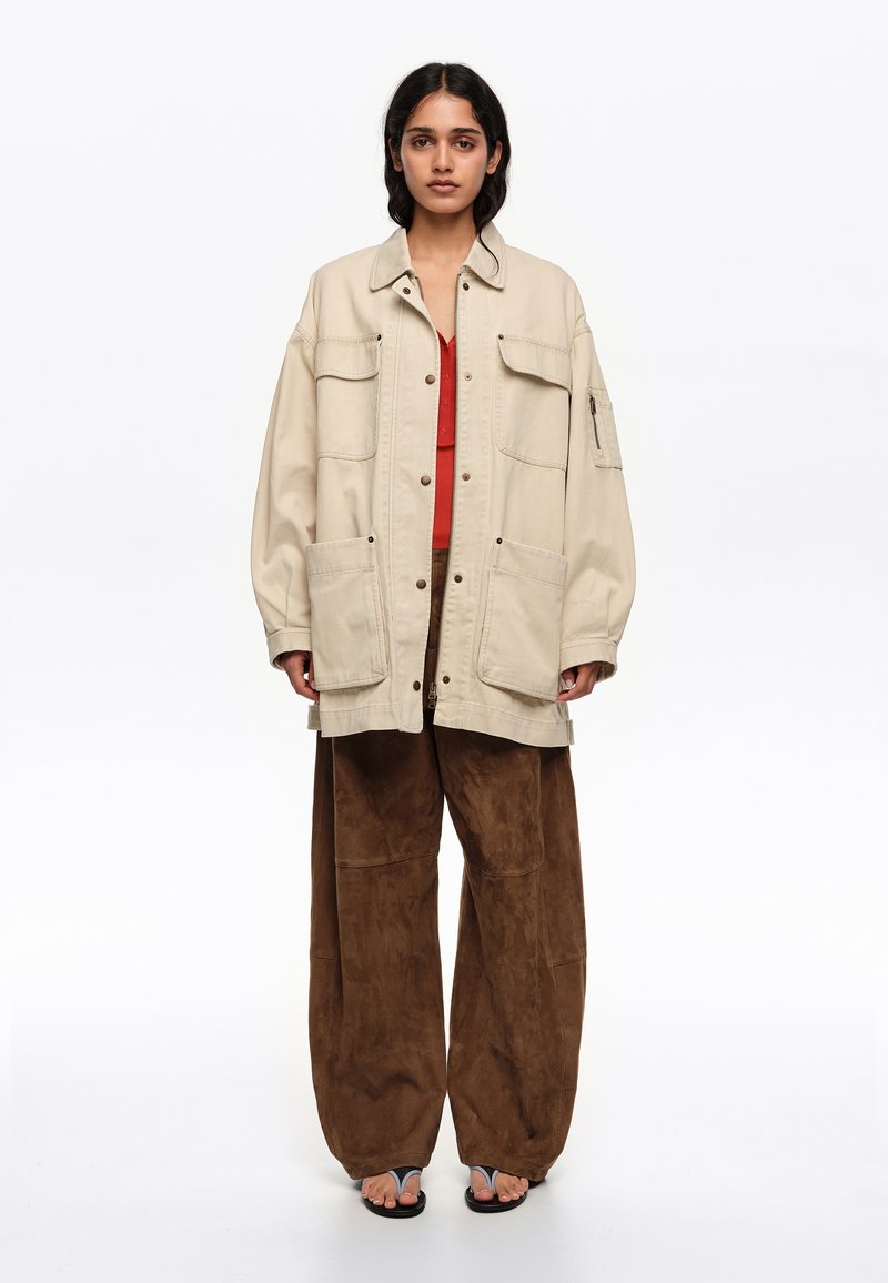 Woman wearing oversized beige jacket, red top, wide brown pants, and black sandals, standing against a plain white background.