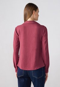 Langermet skjorte i burgunderrød, med krage, teksturert stoff og rundt nederkant, parret med mørkeblå jeans.