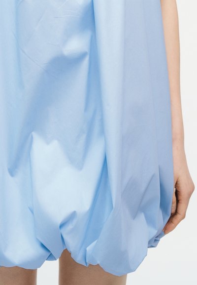 Robe bleu clair à ourlet ballon portée par une personne avec le bras droit et la main visibles sur un fond uni.