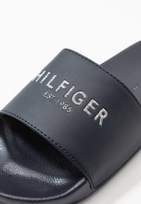 Marinblå slidertöja gjord av slät syntetisk material, med silverbroderad text "HILFIGER EST.1985" på remmen.