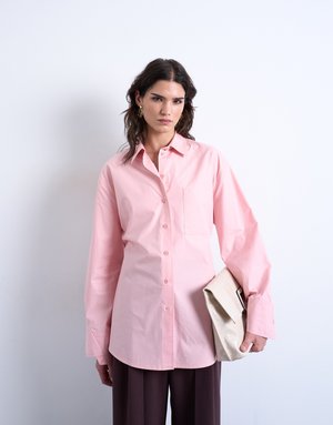 Woman with dark hair wearing oversized pink button-up shirt and dark pants holding a large beige clutch bag against white background.