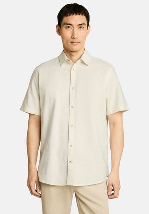 Mann mit kurzen dunklen Haaren, der ein hellbeiges kurzärmeliges Hemd mit Knöpfen und beige Hosen vor einem einfarbigen Hintergrund trägt.