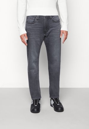 Homme portant un jean gris foncé à jambes droites, une chemise blanche manches longues et des baskets noires avec des accents blancs, debout sur un sol uni.