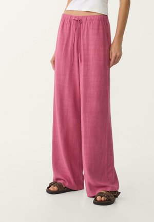 Persoon die losse roze broek met trekkoord en bruine sandalen draagt, staand tegen een effen lichte achtergrond.