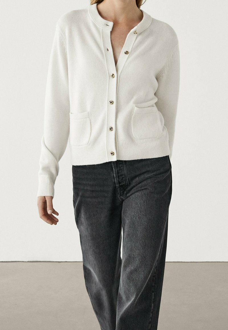 Valkoinen neuletakki, jossa on pyöreä kaula-aukko, ribatut hihansuut ja kaksi etutaskua. Takissa on kultaiset napit ja pehmeä neulepinta. Yhdistetty tummiin farkkuihin.