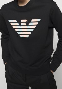 Felpa nera con polsini a coste, con un grande logo di aquila multicolore a strisce orizzontali con accenti rosa pastello e azzurro.