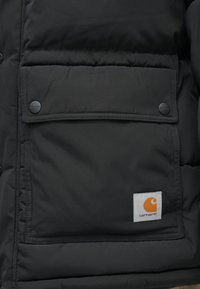 Fekete puffdzseki, nagy elülső zsebbel, fekete snapped gombokkal és egy kiemelkedő narancssárga logóval fehér háttéren.