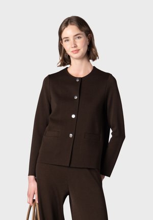 Femme portant une veste boutonnée marron foncé à manches longues et un pantalon assorti, tenant un sac à main beige, debout devant un fond uni.