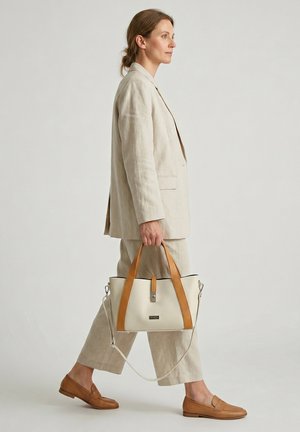 Kvinde i beige jakkesæt går, bærende en creme- og tanfarvet håndtaske, iført tan loafers mod en ensartet lys baggrund.