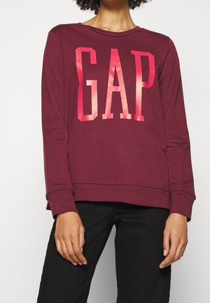 Personne portant un sweat-shirt à manches longues bordeaux avec un grand logo rouge « GAP » et un pantalon noir, debout devant un fond clair uni.