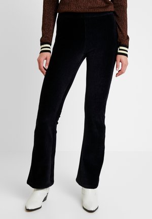 Woman wearing black corduroy flared pants, brown textured sweater with striped cuffs, and white ankle boots against a plain background.